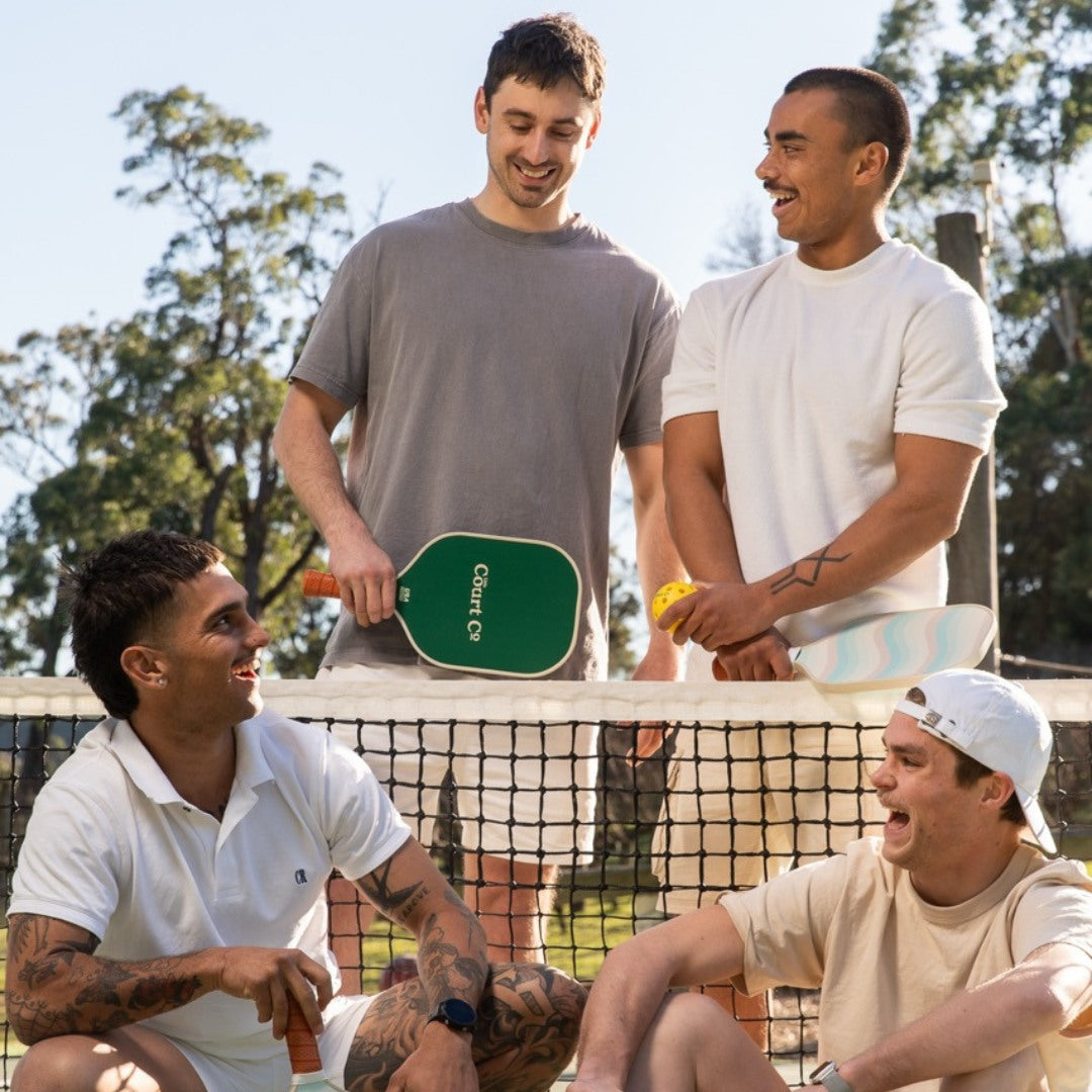 Four men on a pickleball court with pickleball paddles and pickleball equipment, smiling and laughing.