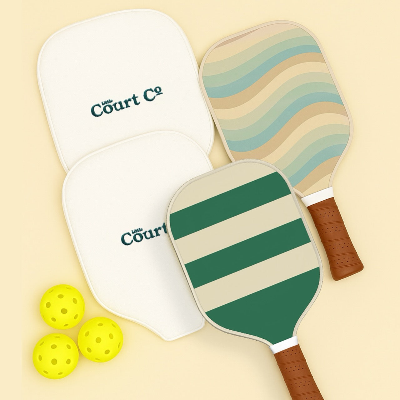 Two paddleball paddles with striped design and a white paddle with 'Little Court Co' branding, accompanied by two yellow balls on a beige background.