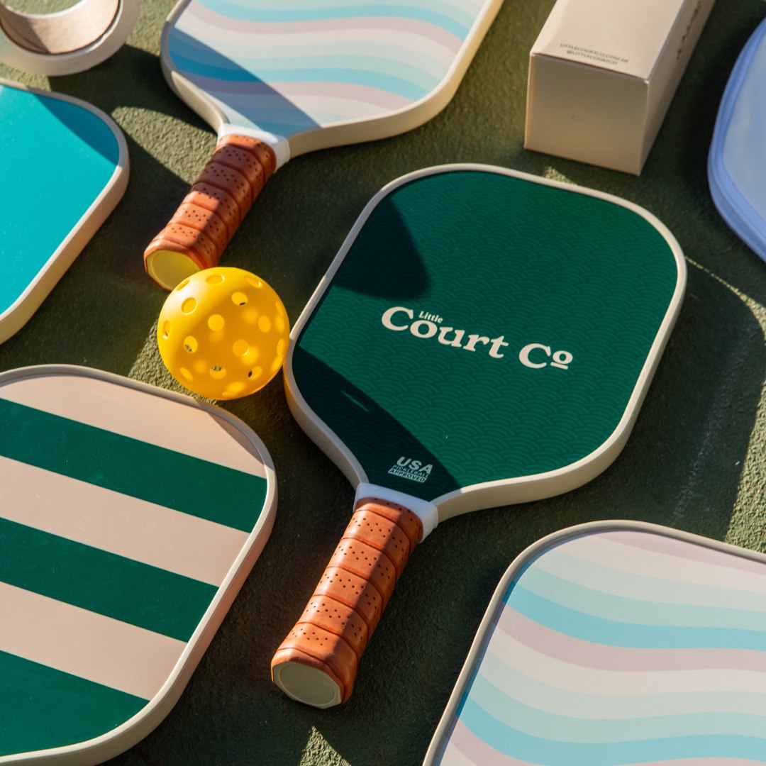 Pickleball paddles with colourful designs on a green surface, featuring 'Little Court Co' branding.